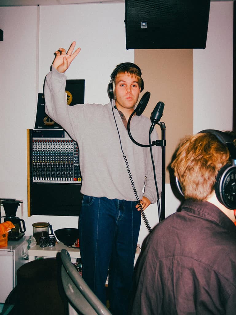 En mann i beige poloskjorte med headset på hodet står i et studio og spiller inn musikk. En mann som sitter ved en pult skimtes i forgrunnen.